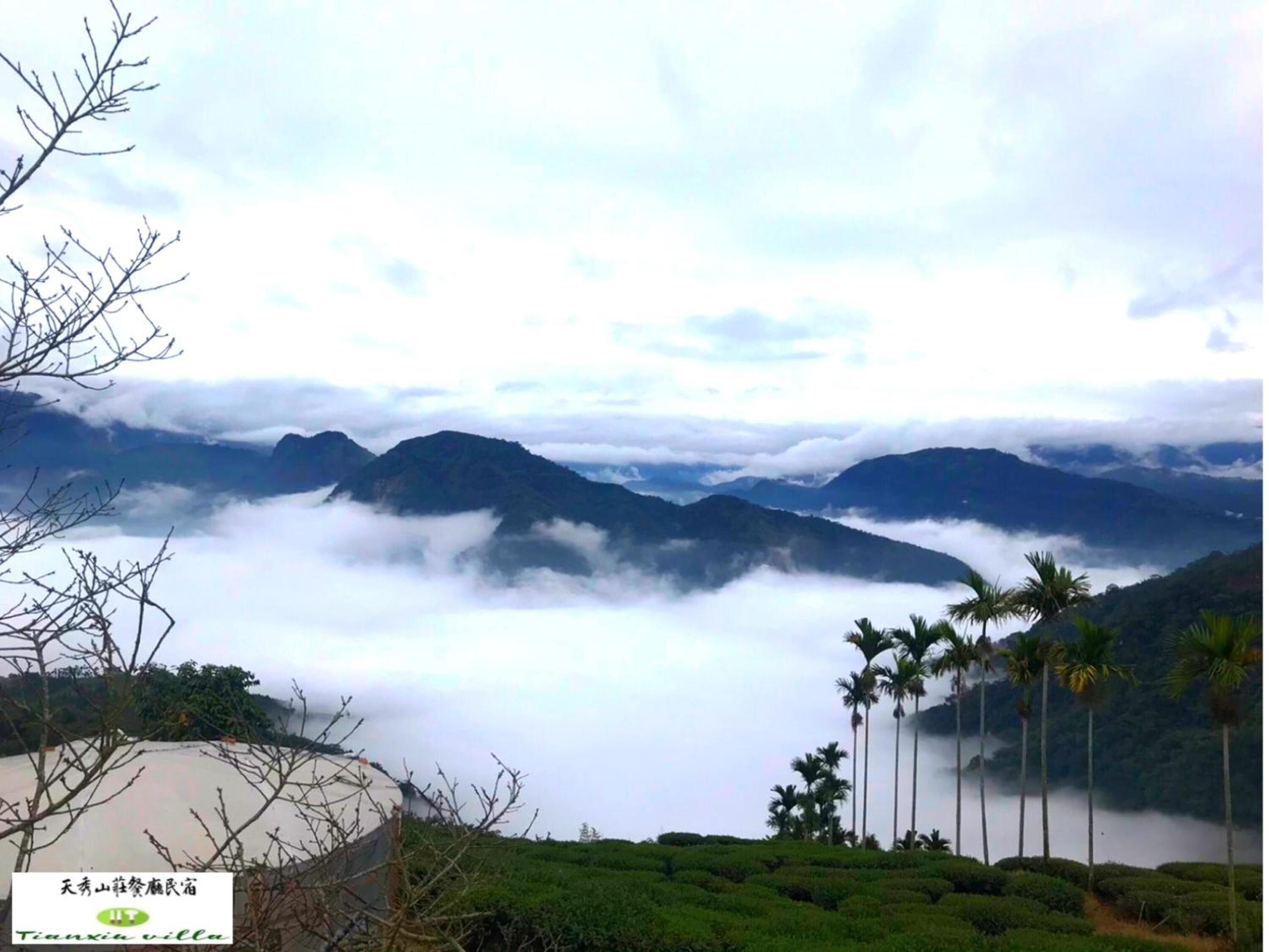 Paisaje montañoso con vista a un valle y un cuerpo de agua. Palmeras y un edificio en primer plano.