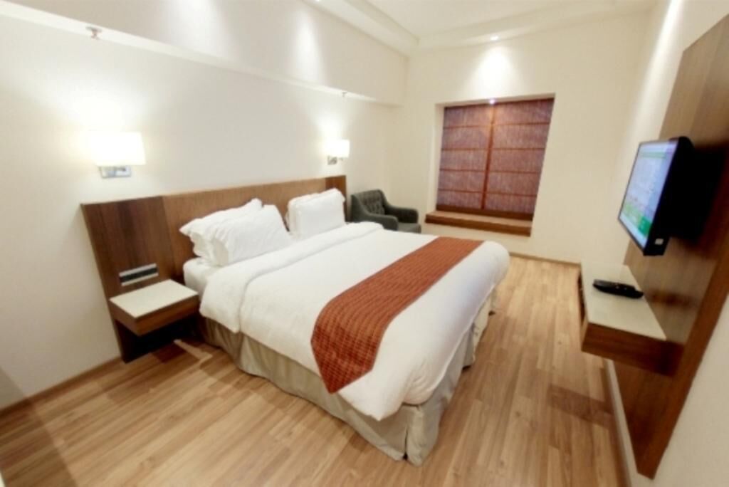 Habitación de hotel con una cama grande, una mesita de noche, una silla, un televisor y una ventana con persianas.