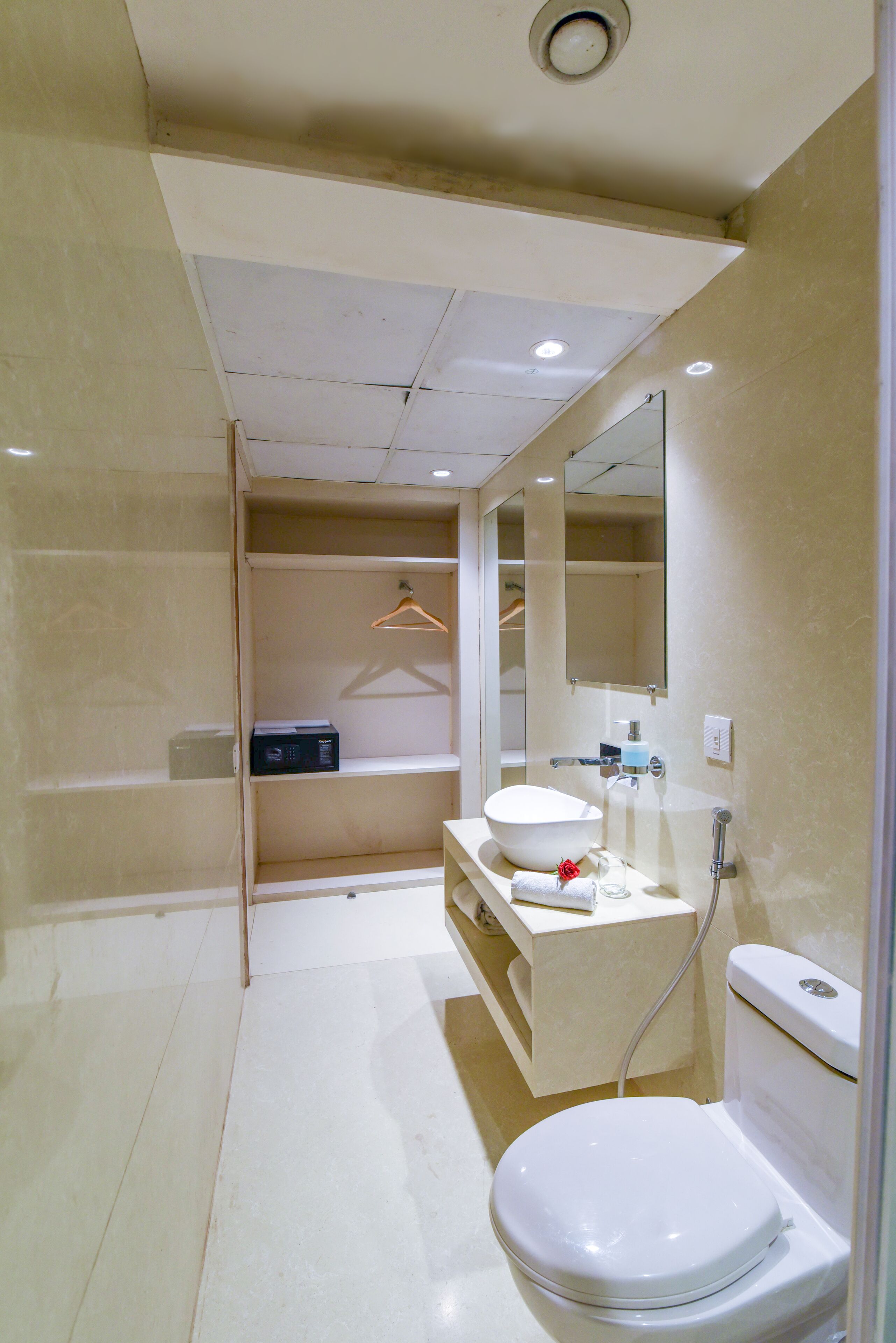 Un baño moderno con lavamanos, espejo y retrete blancos. Se ve un perchero y un estante pequeño con artículos de aseo.