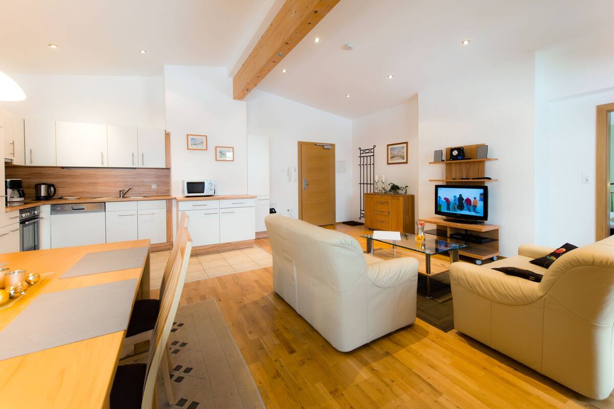 A modern kitchen with white cabinets, a wooden countertop, and a dining area with a table and chairs. There is a living room with a sofa and a television.