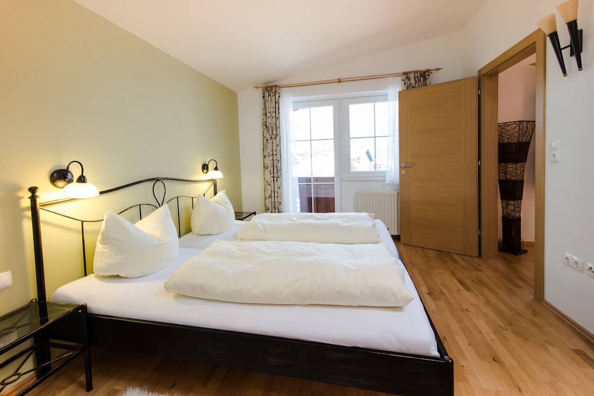 A bedroom with a bed, bedside lamps, a wooden floor, a window with curtains, and a door.