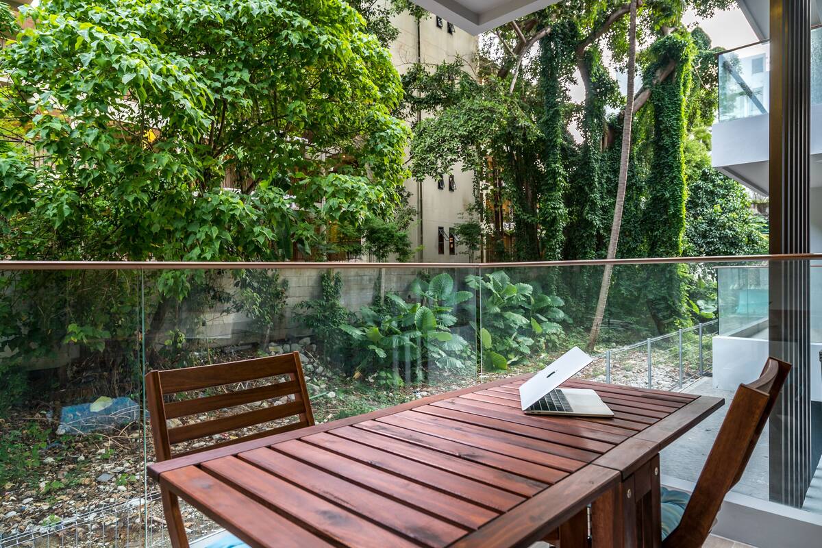 A wooden table with a laptop on it, surrounded by chairs, in a balcony with a glass railing overlooking a garden with various plants and trees.
