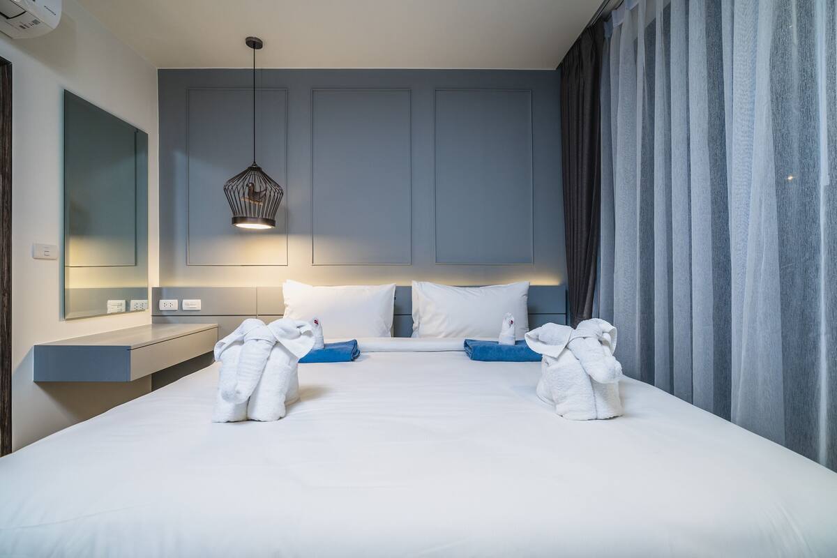 A modern hotel room with a large bed, a bedside table, a hanging light fixture, and a large window with curtains.