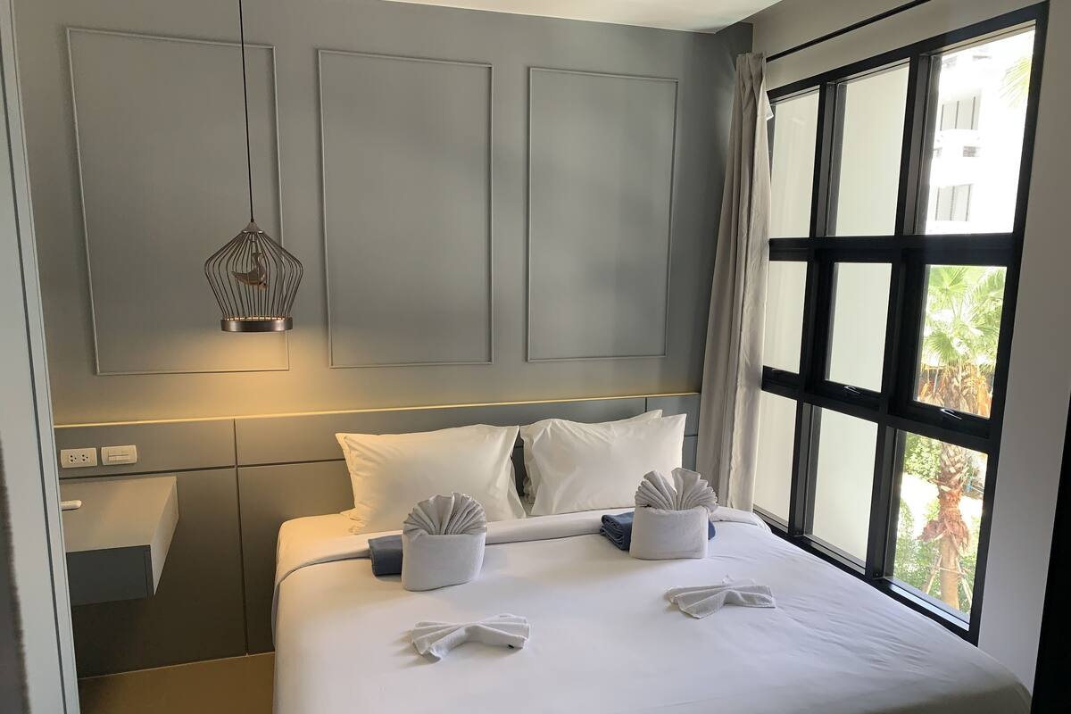 A neatly made bed with white linens, a bedside table with a lamp, and a window with a view of palm trees.