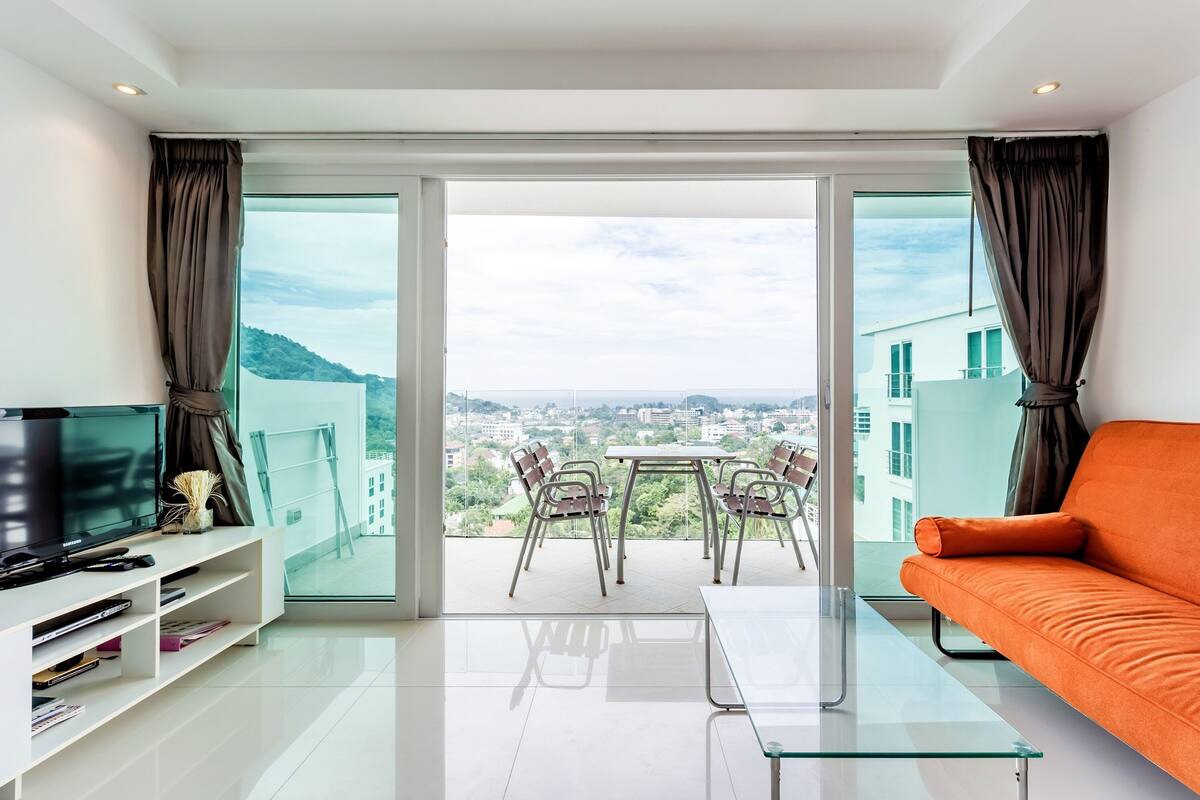 A modern living room with a glass table, a sofa, a TV, and a balcony with a city view.