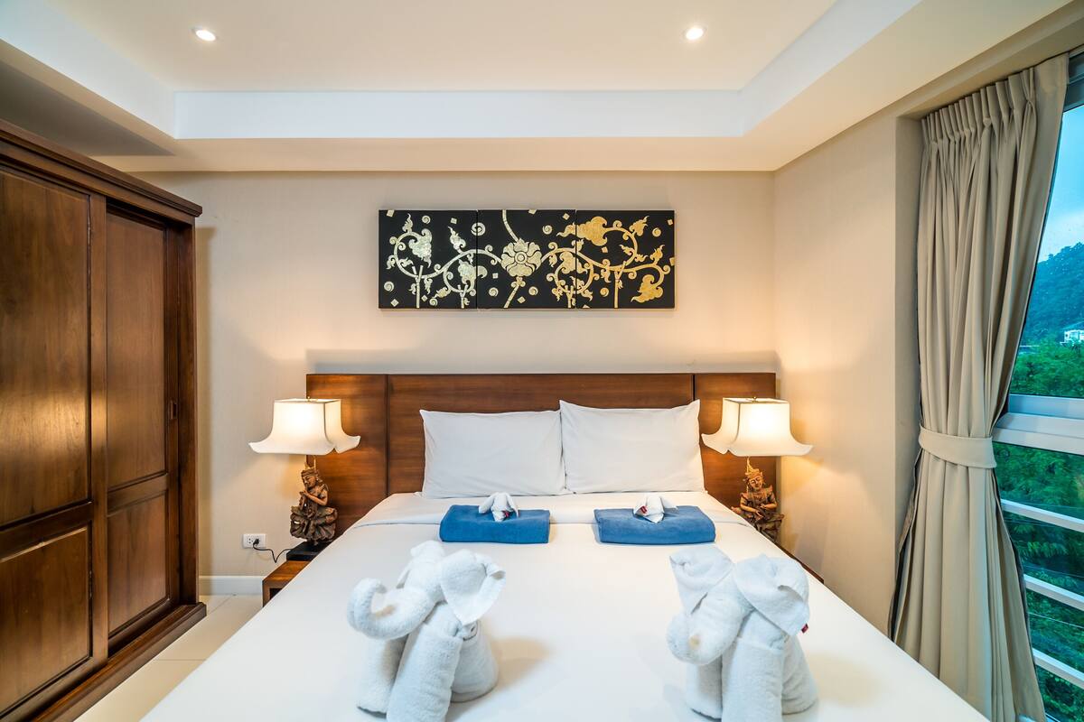 A hotel room with a large bed, two bedside lamps, a wooden headboard, a painting on the wall, and a view of greenery through the window.