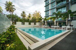 A swimming pool with lounge chairs and umbrellas, surrounded by palm trees and a modern building with balconies. - Oceanstone Resort by Lofty (Choeng Thale)