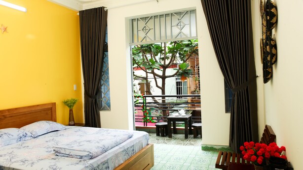 A bedroom with a bed, a small table, and a chair. There is a window with curtains and a view of a tree outside. - PT House (Ho Chi Minh City)