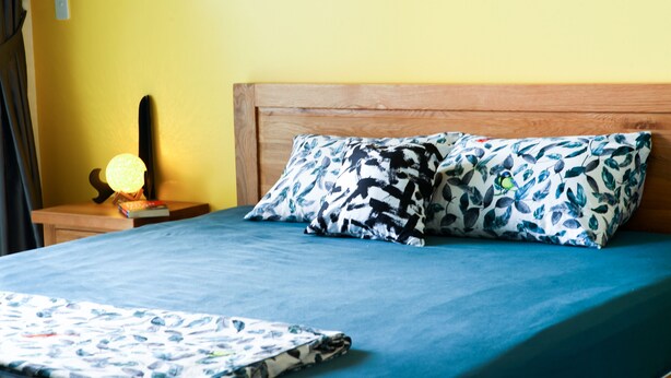 A bed with a blue sheet, two patterned pillows, a wooden headboard, a bedside lamp, and a nightstand with a book and a lamp. - PT House (Ho Chi Minh City)