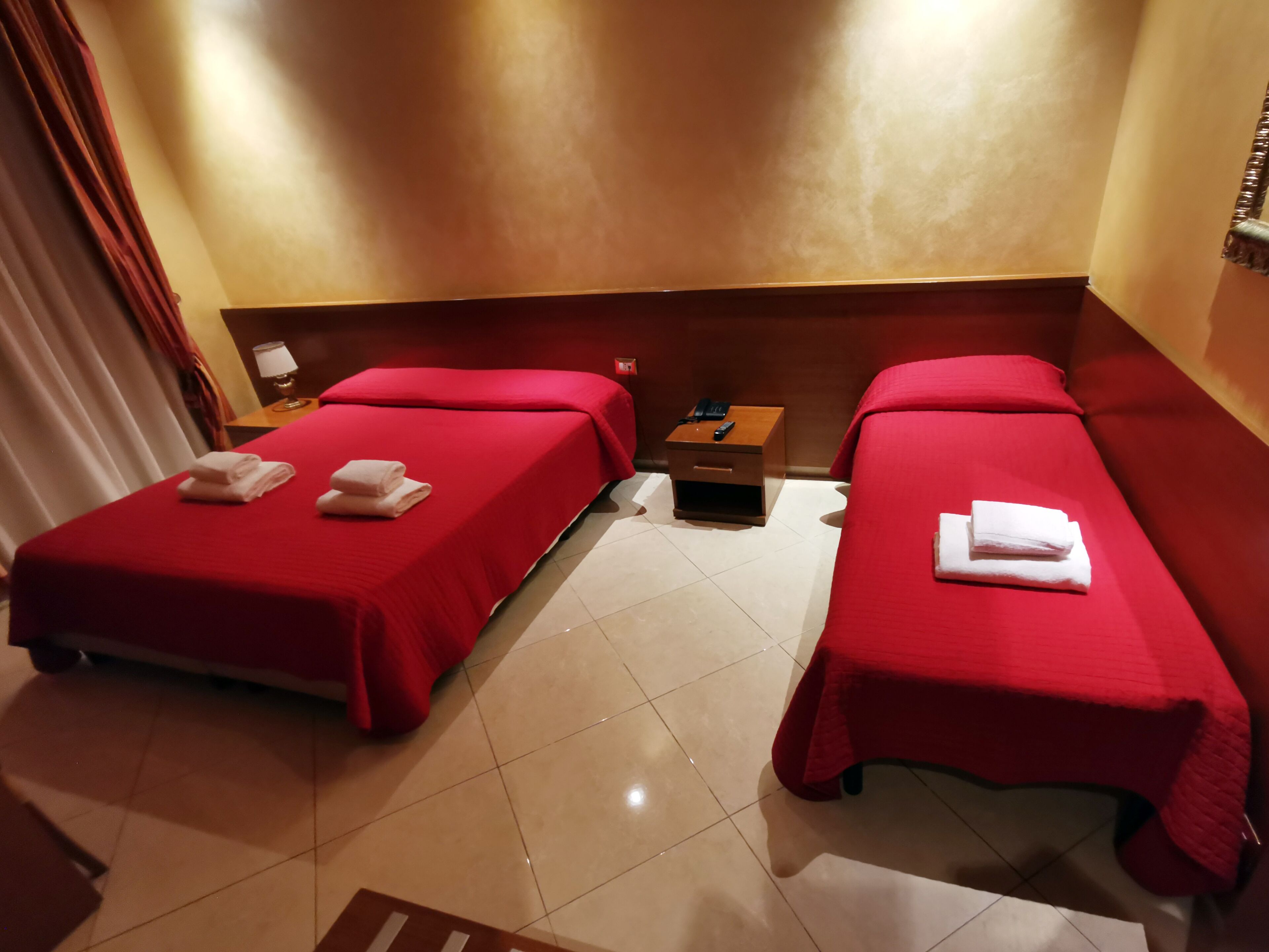 Ein Hotelzimmer mit zwei Betten, jeweils mit roter Bettdecke und weißen Handtüchern. Dazwischen befindet sich ein Nachttisch mit Lampe und Telefon.