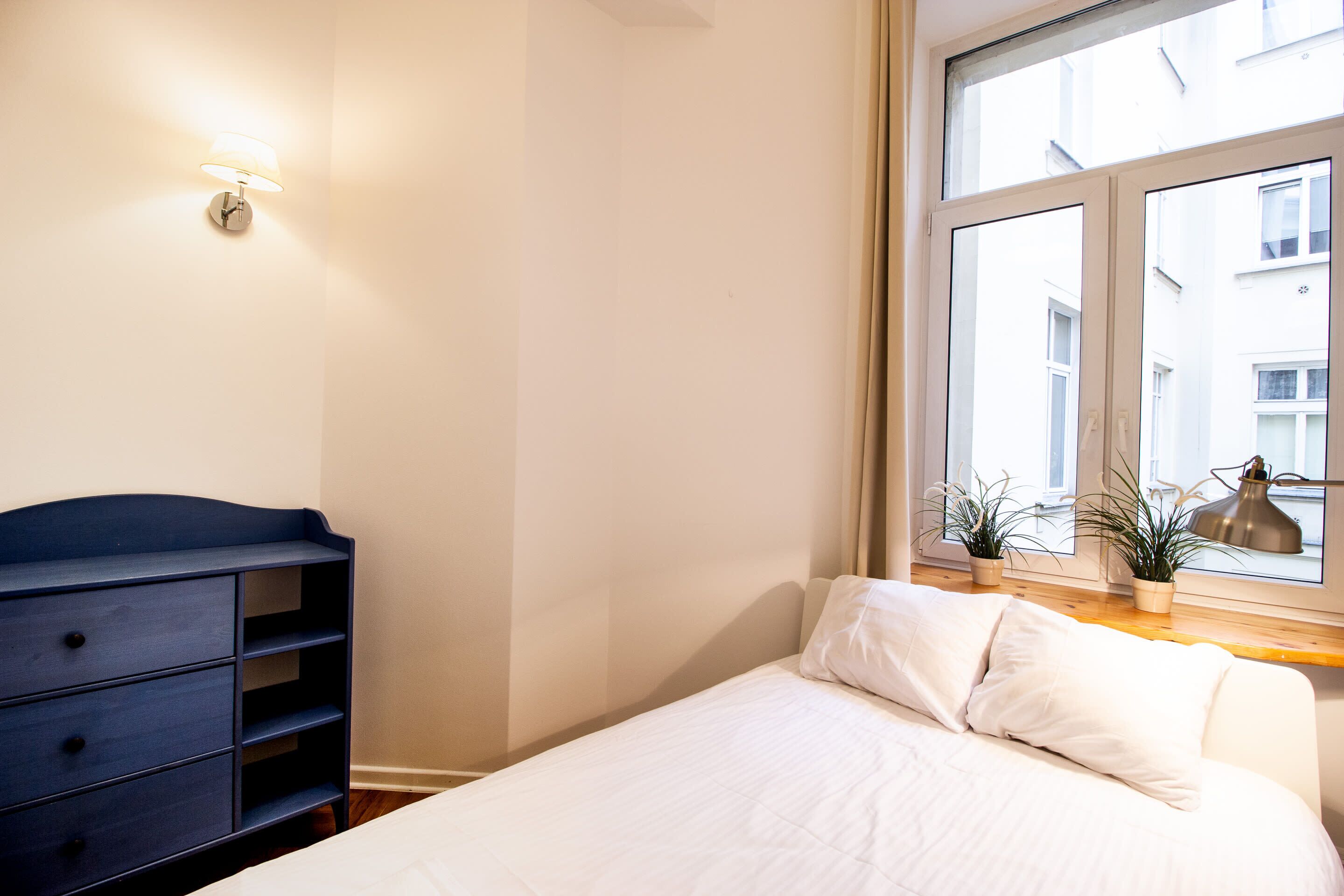 A bedroom with a bed, a dresser, and a window with plants.