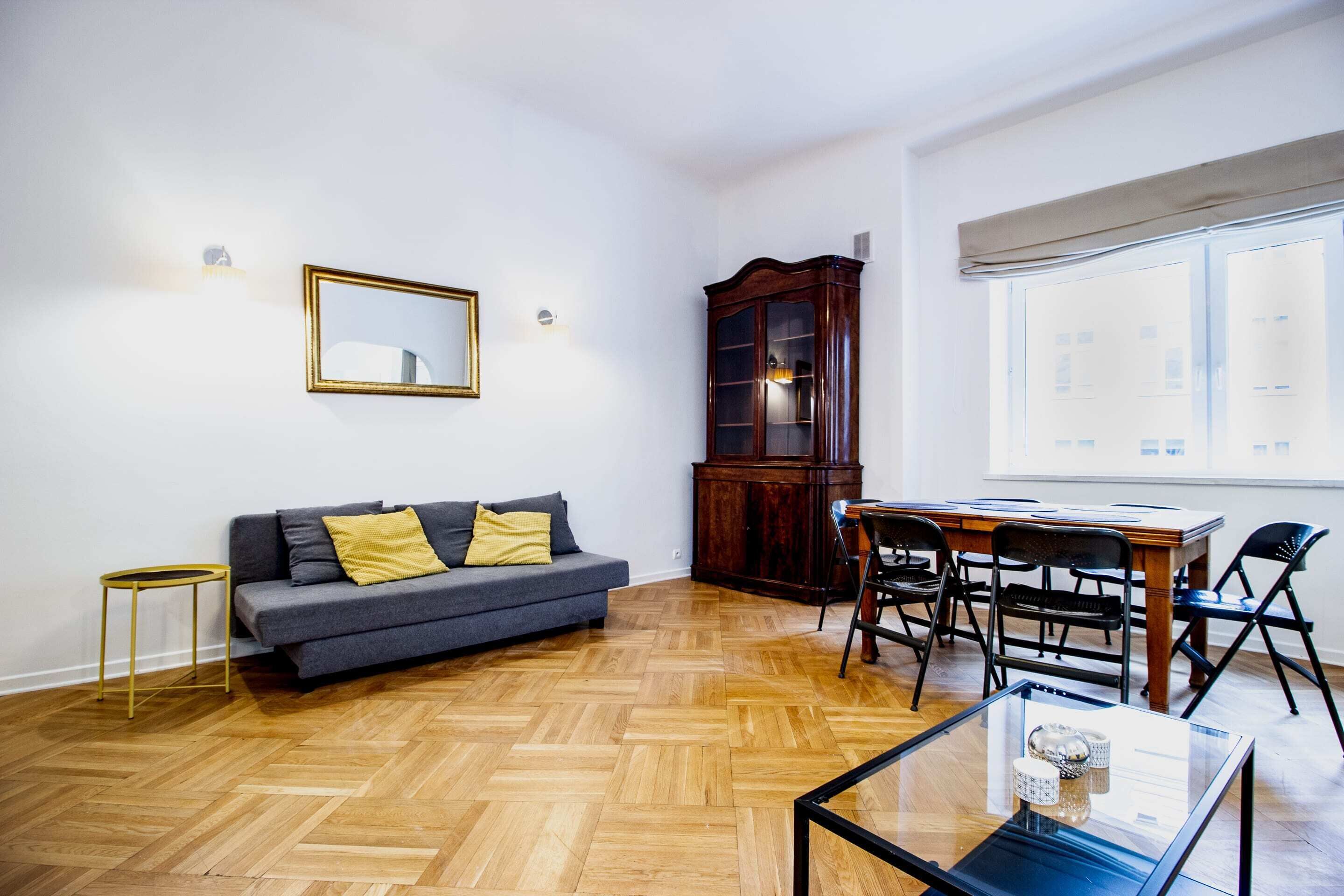 Una sala de estar con un sofá, piso de madera en espiga, una mesa de comedor con sillas, un armario y una ventana con cortinas.