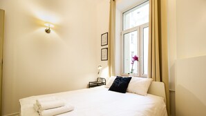 A neatly made bed with white linens and a folded towel, a bedside table with a lamp, and a window with curtains. - Kopernika Srodmiescie Apartment (Warsaw)