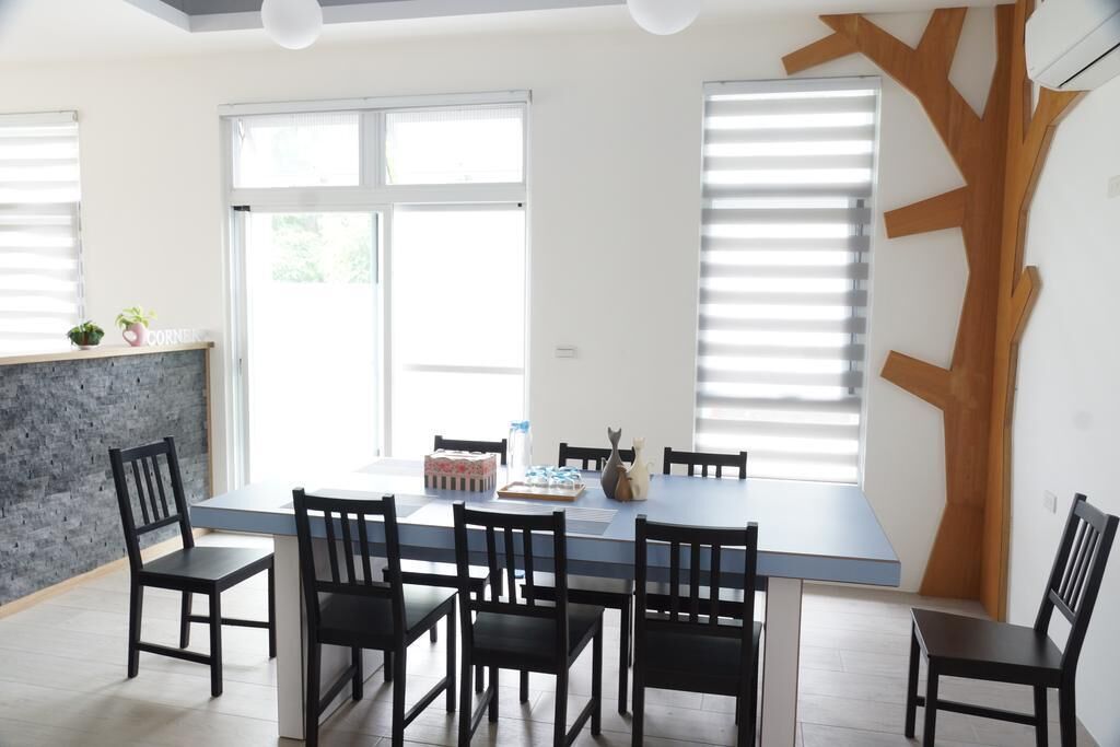 A modern dining area with a long table and six chairs, large windows with blinds, and a unique wooden wall design.
