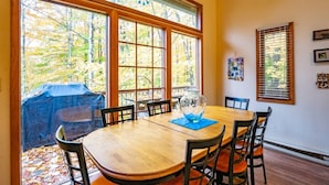 Un comedor con una mesa grande y sillas, ventanas amplias con vistas a árboles, y una zona de estar exterior cubierta.