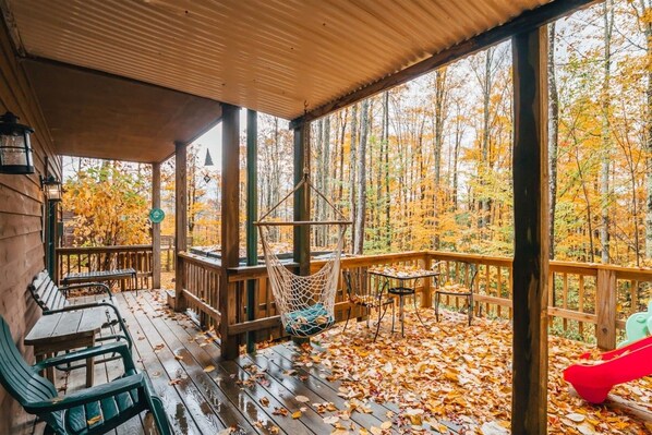A covered outdoor deck with wooden furniture, a hanging chair, and a view of a forest in autumn.