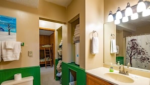 Baño con inodoro, lavamanos y espejo. Una pared verde y un cuadro de un árbol.