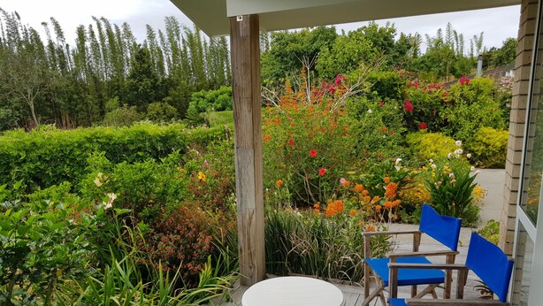 Una terraza de madera con una mesa redonda y sillas azules, rodeada de abundante vegetación y plantas con flores.