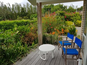 A wooden deck with a round table and blue chairs, surrounded by lush greenery and flowering plants. - Rural retreat minutes from the beaches of Waipu (Waipu)