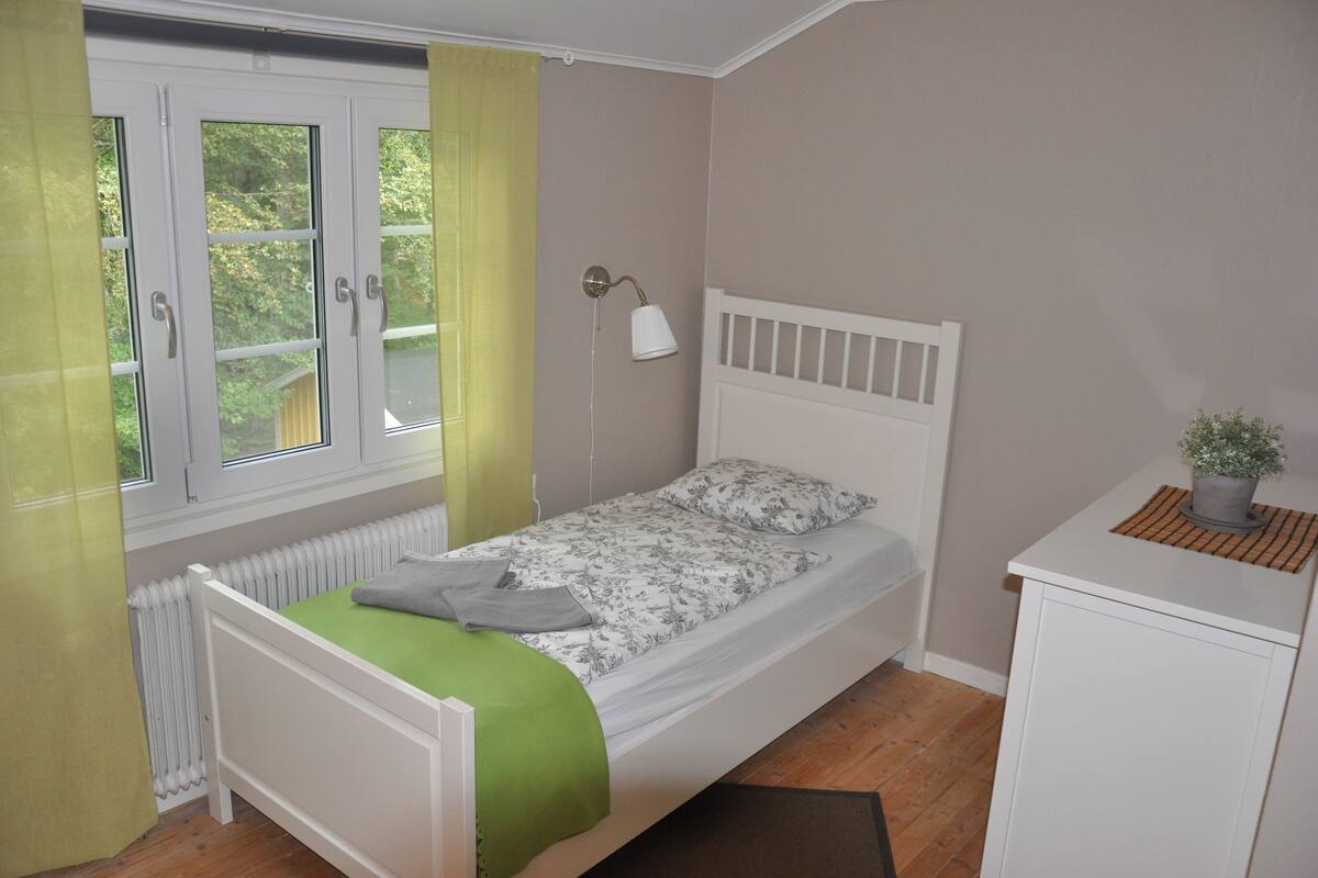 Une chambre avec un lit simple, une tête de lit blanche, une couverture verte, une petite table de chevet avec un pot de plante et une fenêtre avec des rideaux.