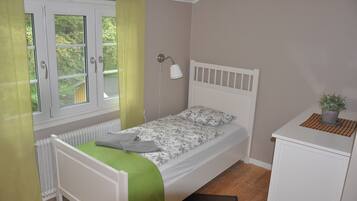 Habitación individual con una cama y cabecera blanca, una manta verde, una mesita de noche con una planta en maceta y una ventana con cortinas.