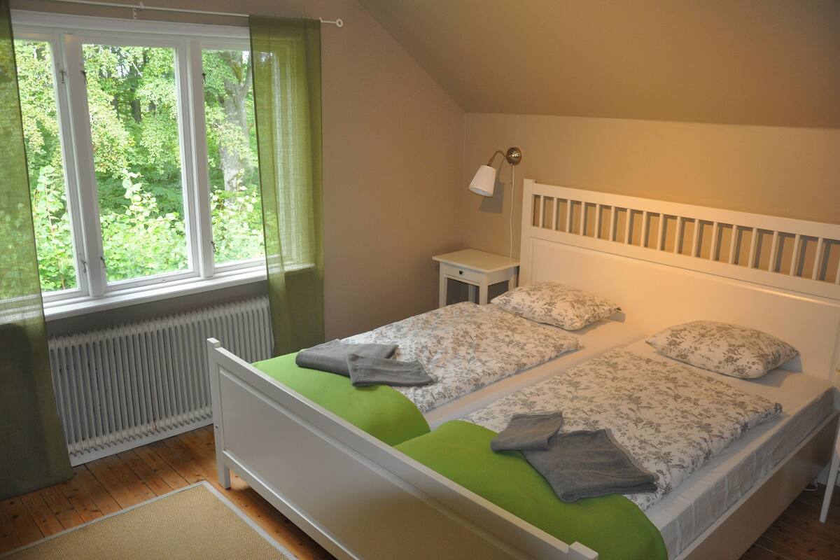 Une chambre avec deux lits simples, une table de chevet, une lampe et une fenêtre donnant sur un paysage verdoyant.