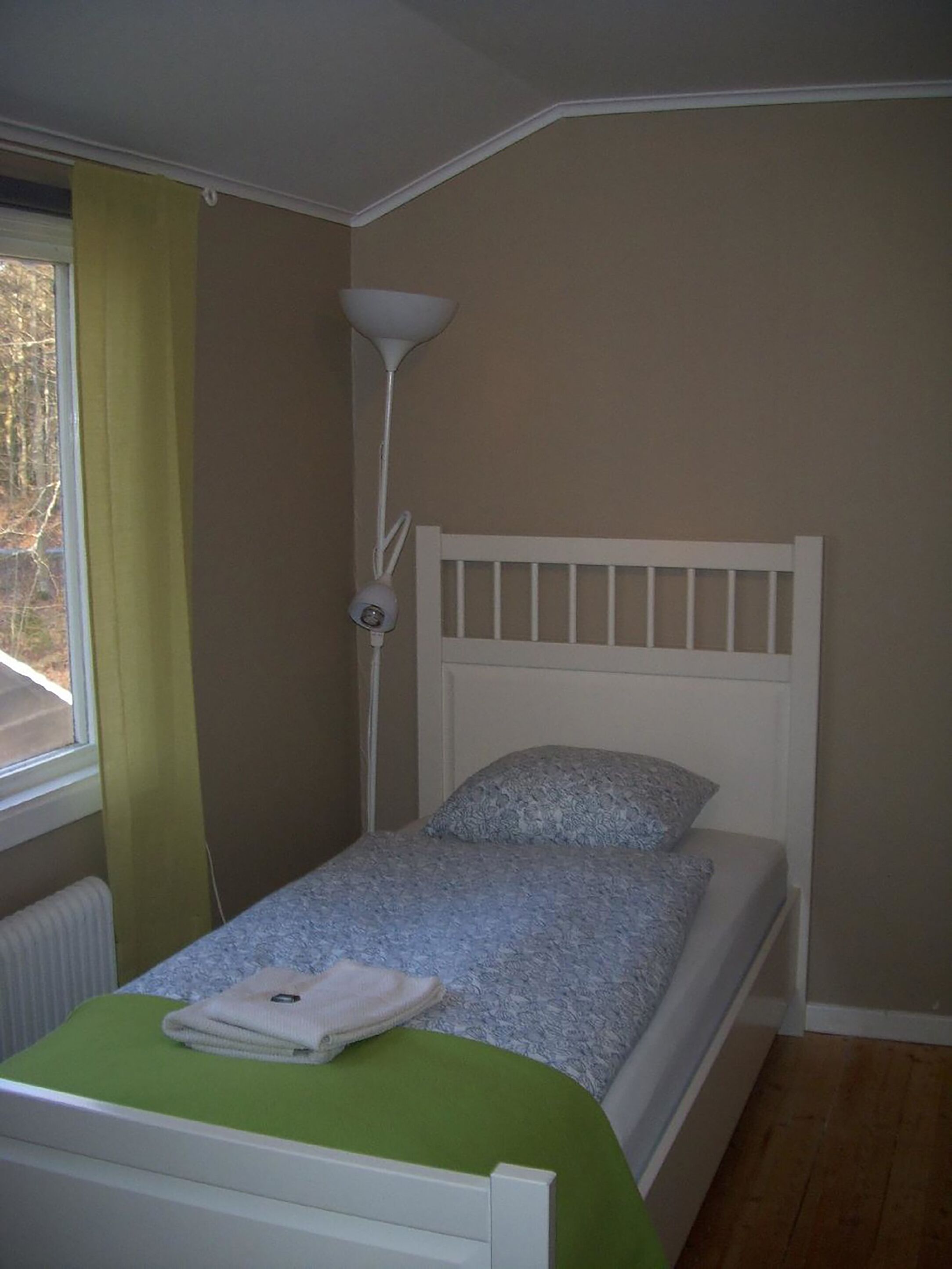 Ein kleines Zimmer mit einem Einzelbett, einem weißen Kopfstück, einer grünen Tagesdecke und einer Lampe am Bett.