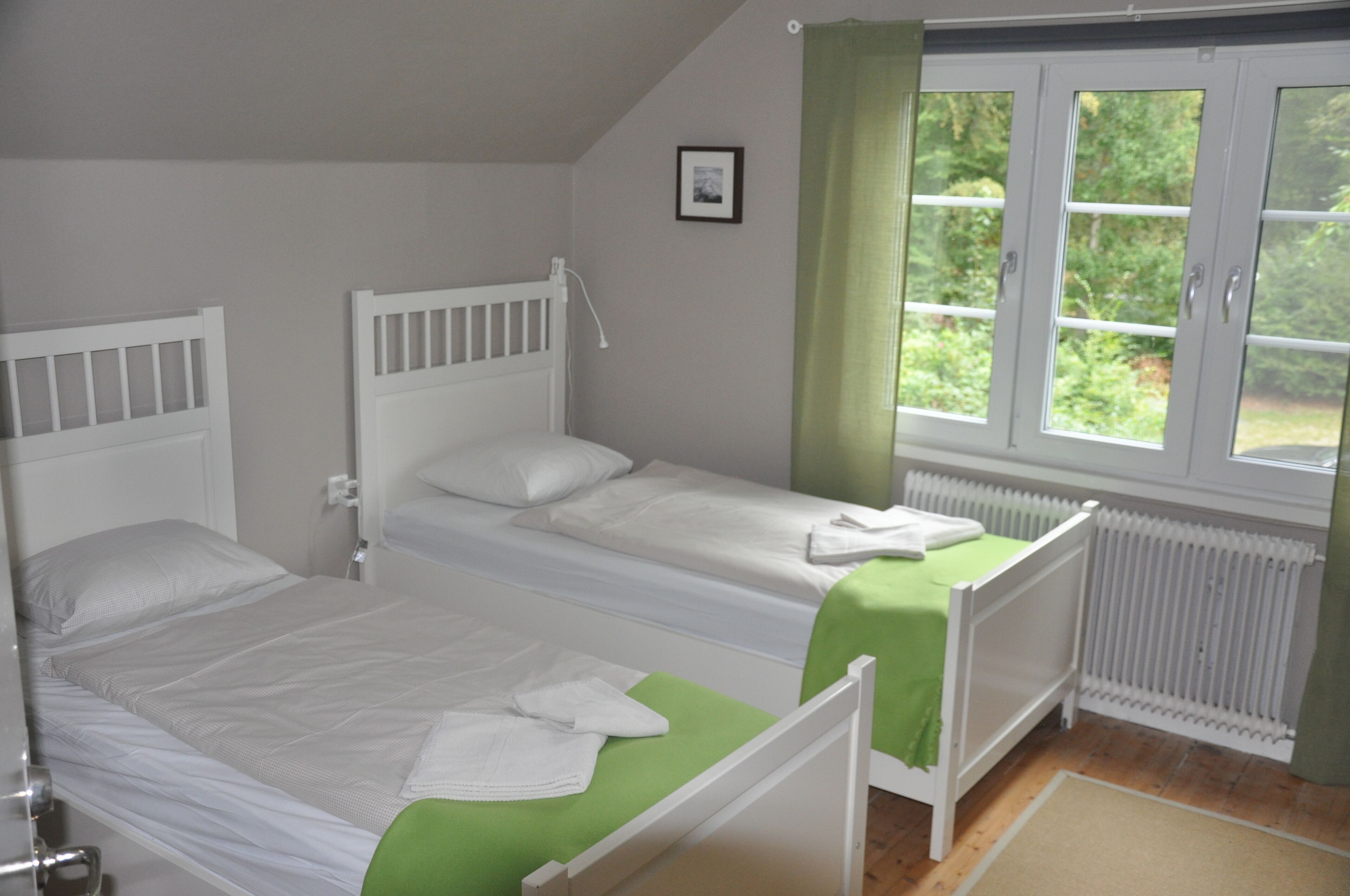 A bedroom with two single beds, a window with curtains, a radiator, and a small framed picture on the wall.