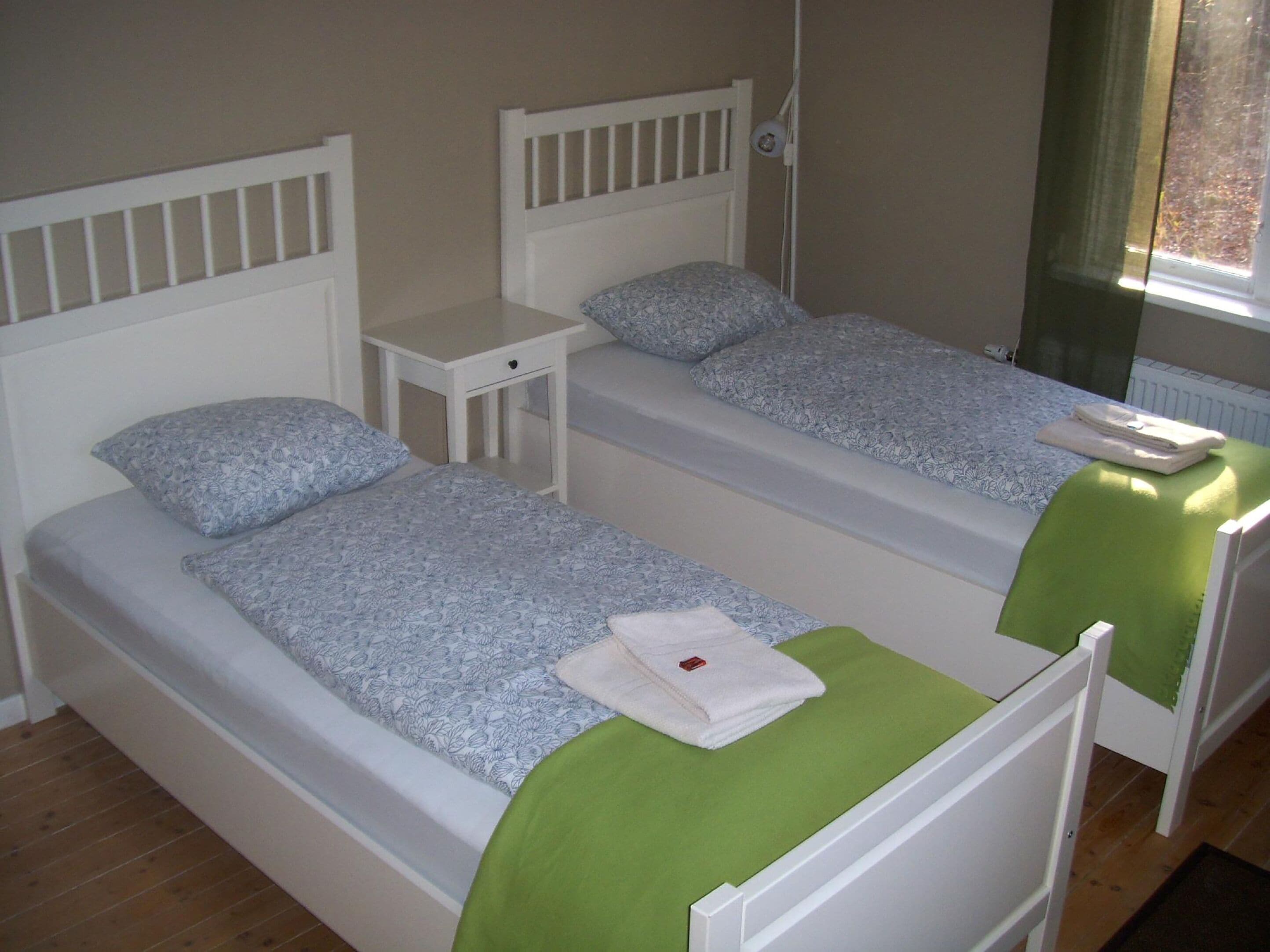 A room with two single beds, each with a white headboard, a small bedside table, and a green blanket on one bed.