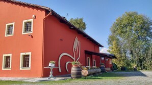 A red building with multiple windows, a large tree in the background, and outdoor potted plants. - Future is Nature Playground (Sala Biellese)