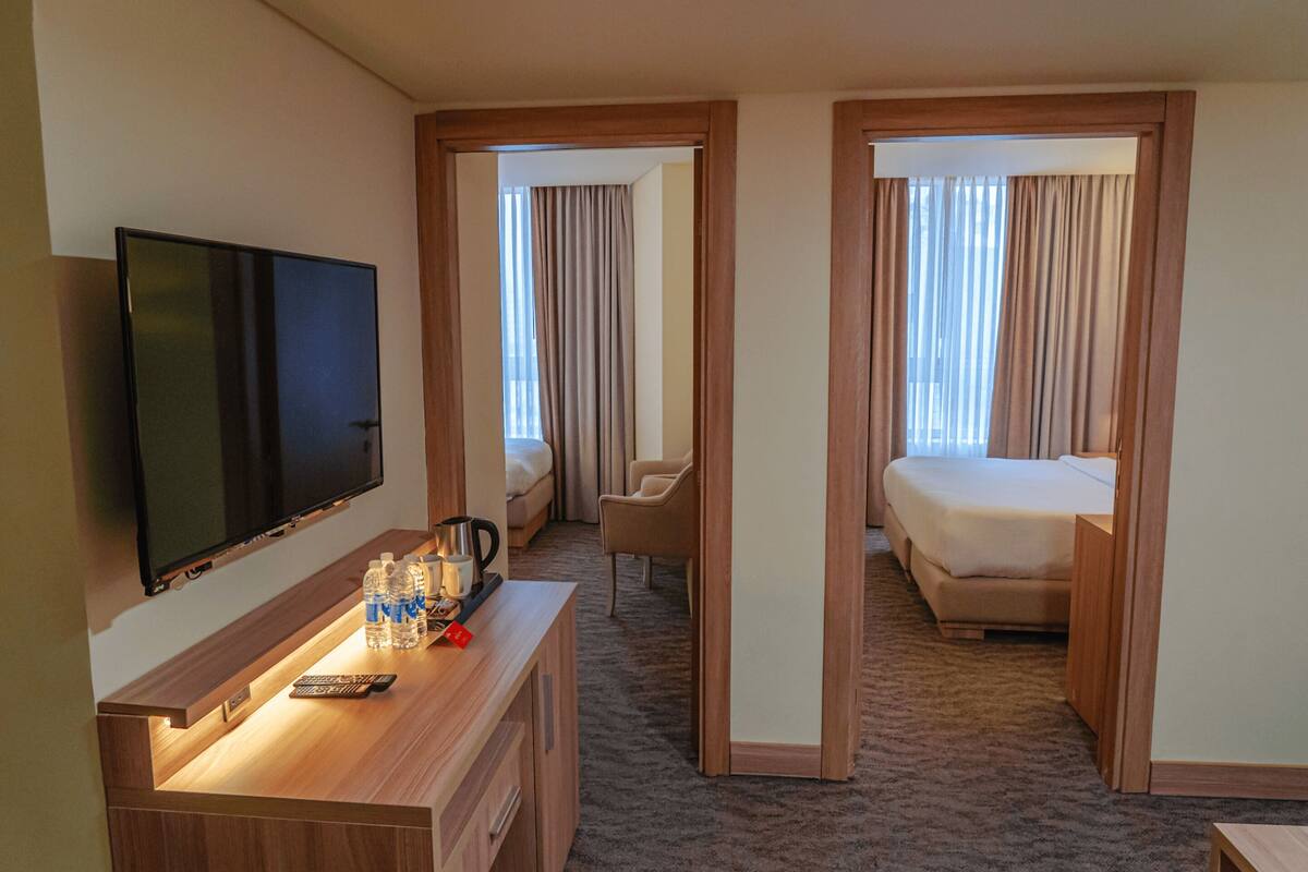 Ein Hotelzimmer mit Flachbildfernseher, Schreibtisch mit Fernbedienung, Wasserflaschen und einer Kaffeemaschine. Im Zimmer befinden sich zwei Betten und ein Sessel.