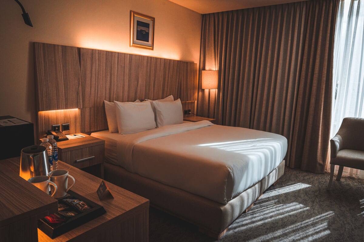 Ein Hotelzimmer mit einem großen Bett, Nachttischlampen, einem hölzernen Kopfteil, einem Schreibtisch mit Kaffeemaschine und einem Sessel.
