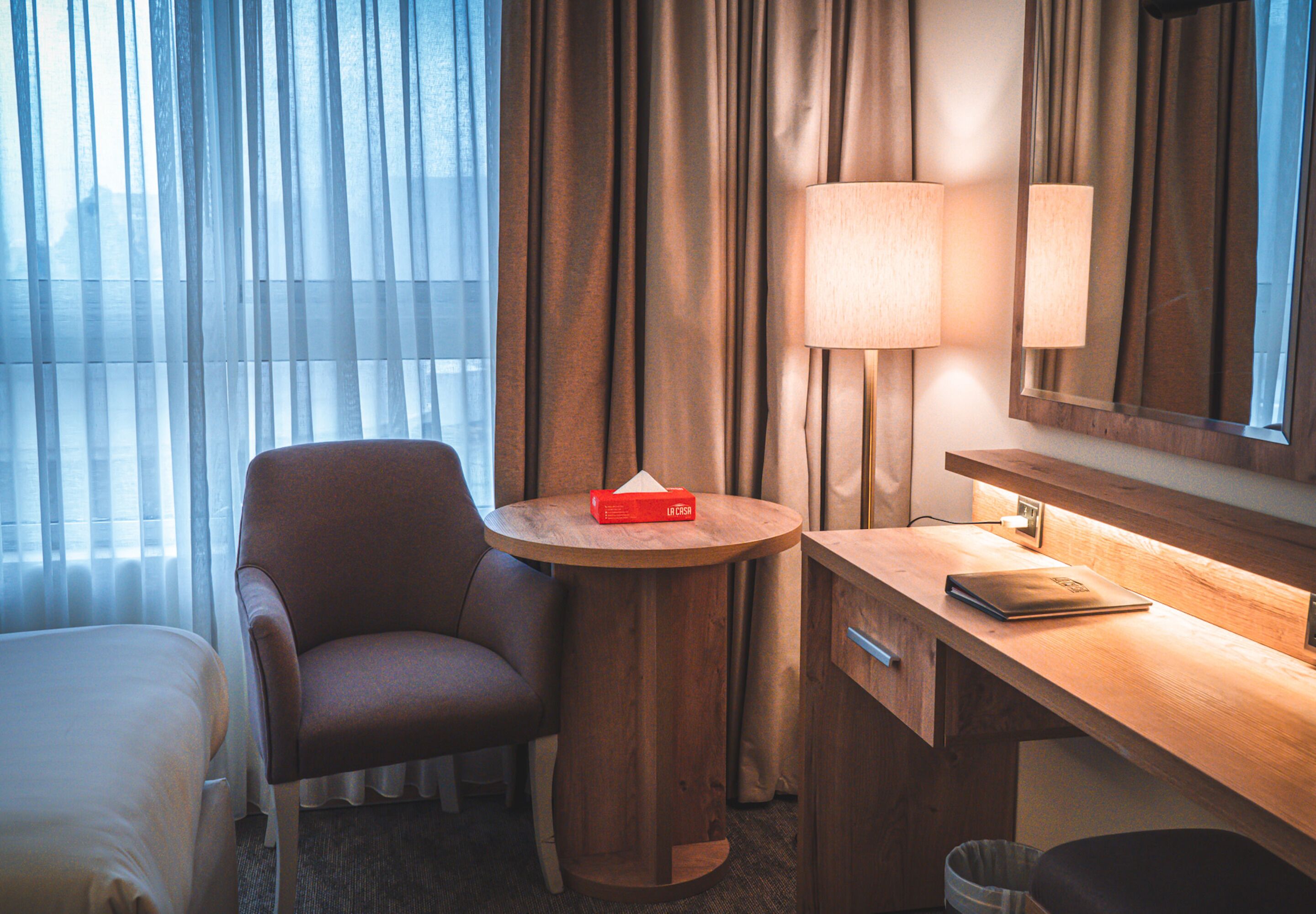 A hotel room with a bed, a chair, a round wooden table with a lamp, a mirror, and a trash bin.