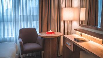 A hotel room with a bed, a chair, a round wooden table with a lamp, a mirror, and a trash bin.