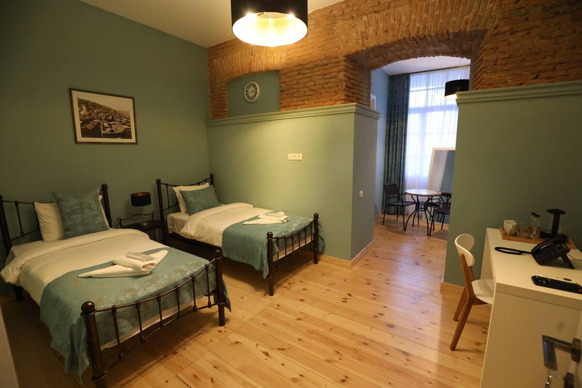 A room with two beds, a desk, a chair, a clock, and a window with curtains.