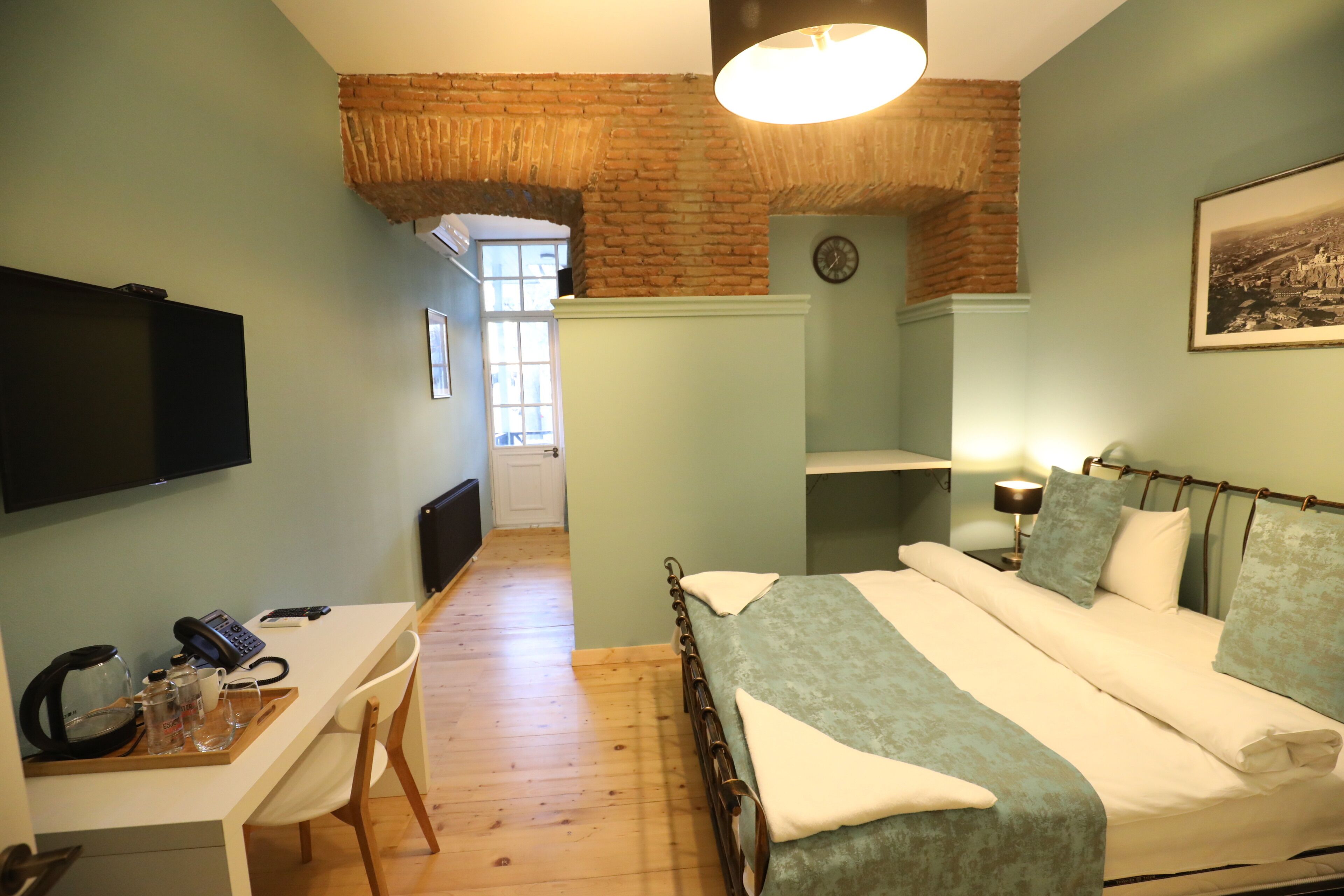 Un dormitorio con una cama, un escritorio con teléfono, gafas, y una tetera, pared de ladrillo, ventana y un reloj en la pared.