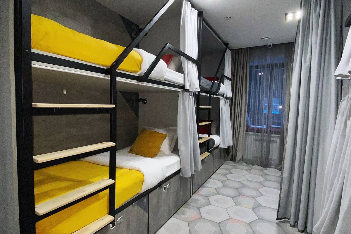 Une chambre avec trois lits superposés, chacun étant équipé d’une couverture jaune et de draps blancs. Le sol de la pièce est recouvert d’un motif géométrique et il y a une fenêtre avec des rideaux.