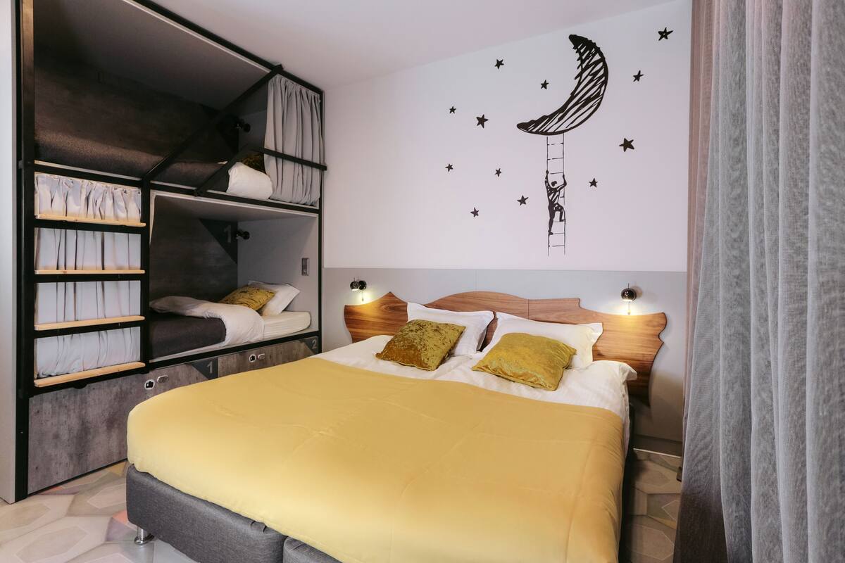 Une chambre avec un lit superposé, un lit recouvert d’une couverture jaune et un motif décoratif représentant la lune et les étoiles sur un mur.