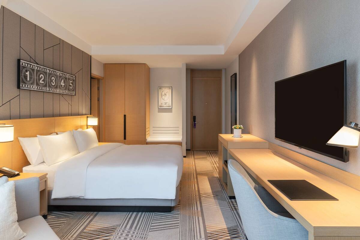 A modern hotel room with a large bed, a bedside table with a lamp, a flat-screen TV mounted on the wall, and a large mirror.