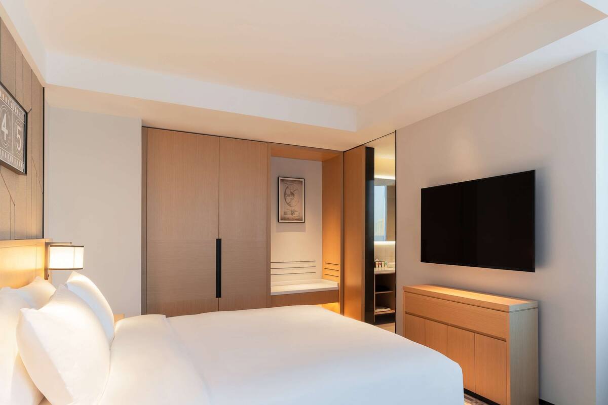 A hotel room with a large bed, a TV mounted on the wall, a wardrobe, and a bedside table with a lamp.