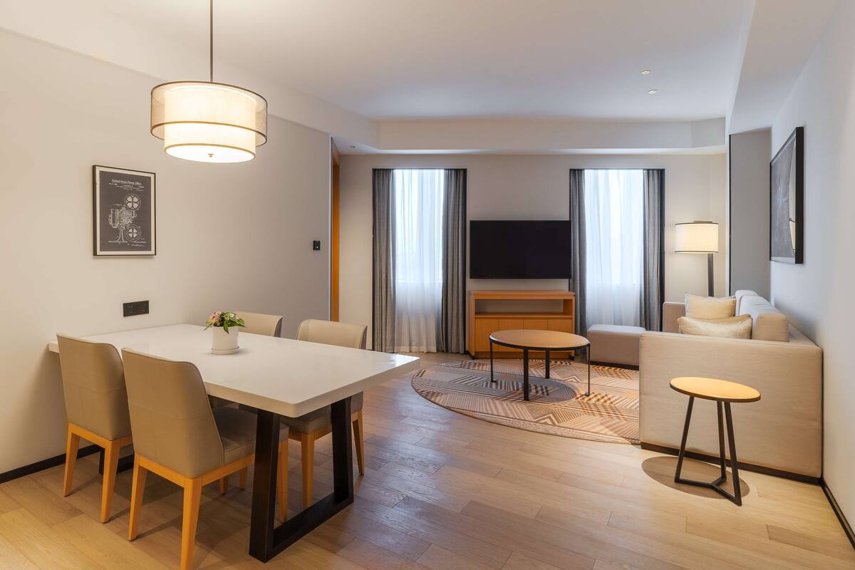 A modern hotel room with a dining area, a sofa, a TV, and a lamp.