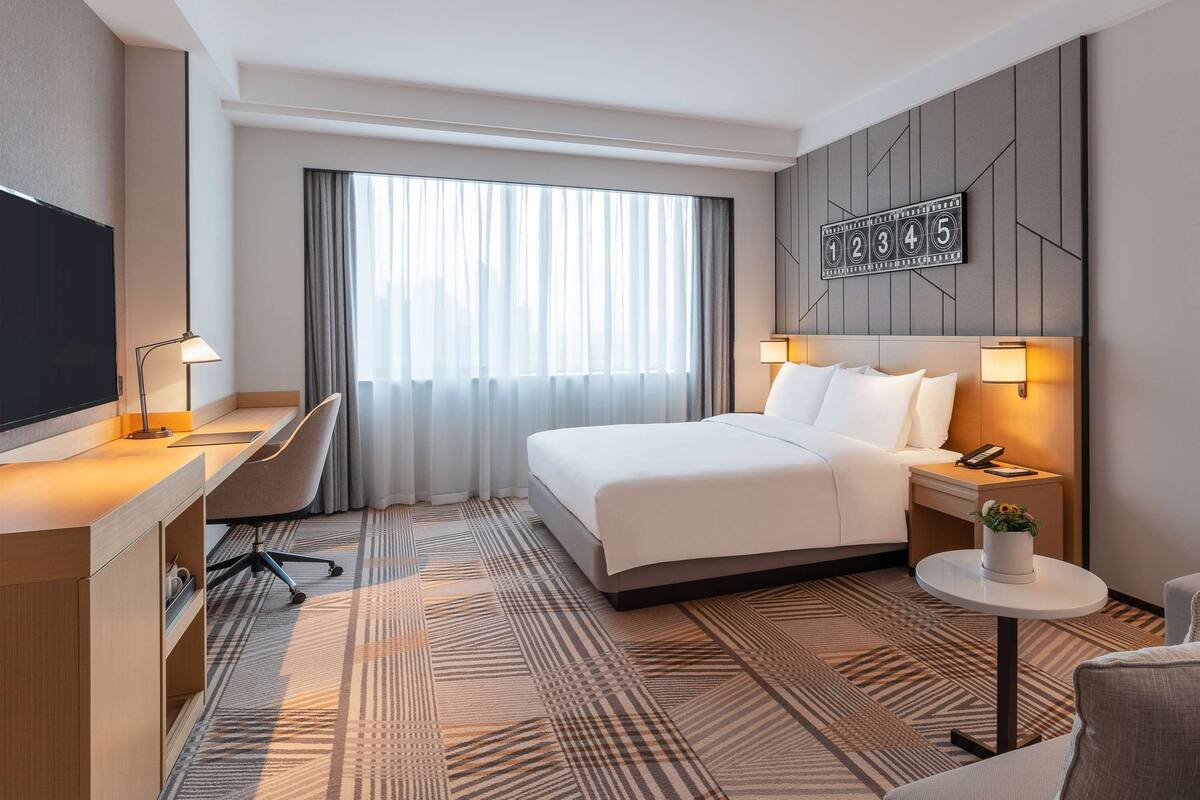 A hotel room with a large bed, a desk with a chair, a TV, a bedside table with a lamp, and a small round table with a plant.