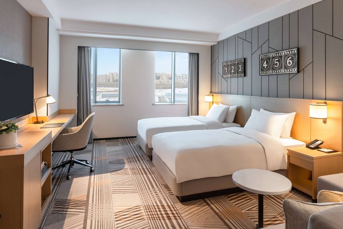A hotel room with two beds, a desk, a chair, a small table, and a TV.