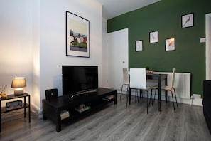 A living room with a flat-screen TV, a dining table with chairs, and a green accent wall.