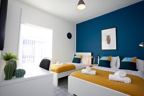 A hotel room with two beds, a desk, a chair, a potted plant, and a window with a view of buildings. - Salford House (Salford)