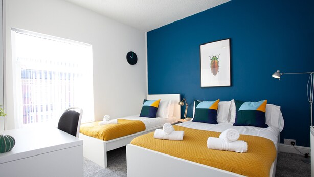 A hotel room with two beds, a desk, a chair, a clock, a lamp, and a window with blinds. - Salford House (Salford)