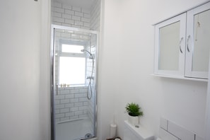 A bathroom with a glass-enclosed shower, white tiled walls, a white cabinet, and a small potted plant.