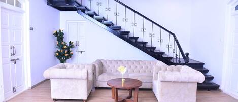 Una sala de estar con un sofá seccional color beige, una mesa de centro redonda de madera y una escalera con barandillas negras.