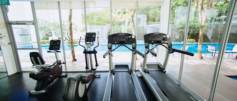 A gym with treadmills, stationary bikes, and a rowing machine, large windows, and a view of a pool outside.