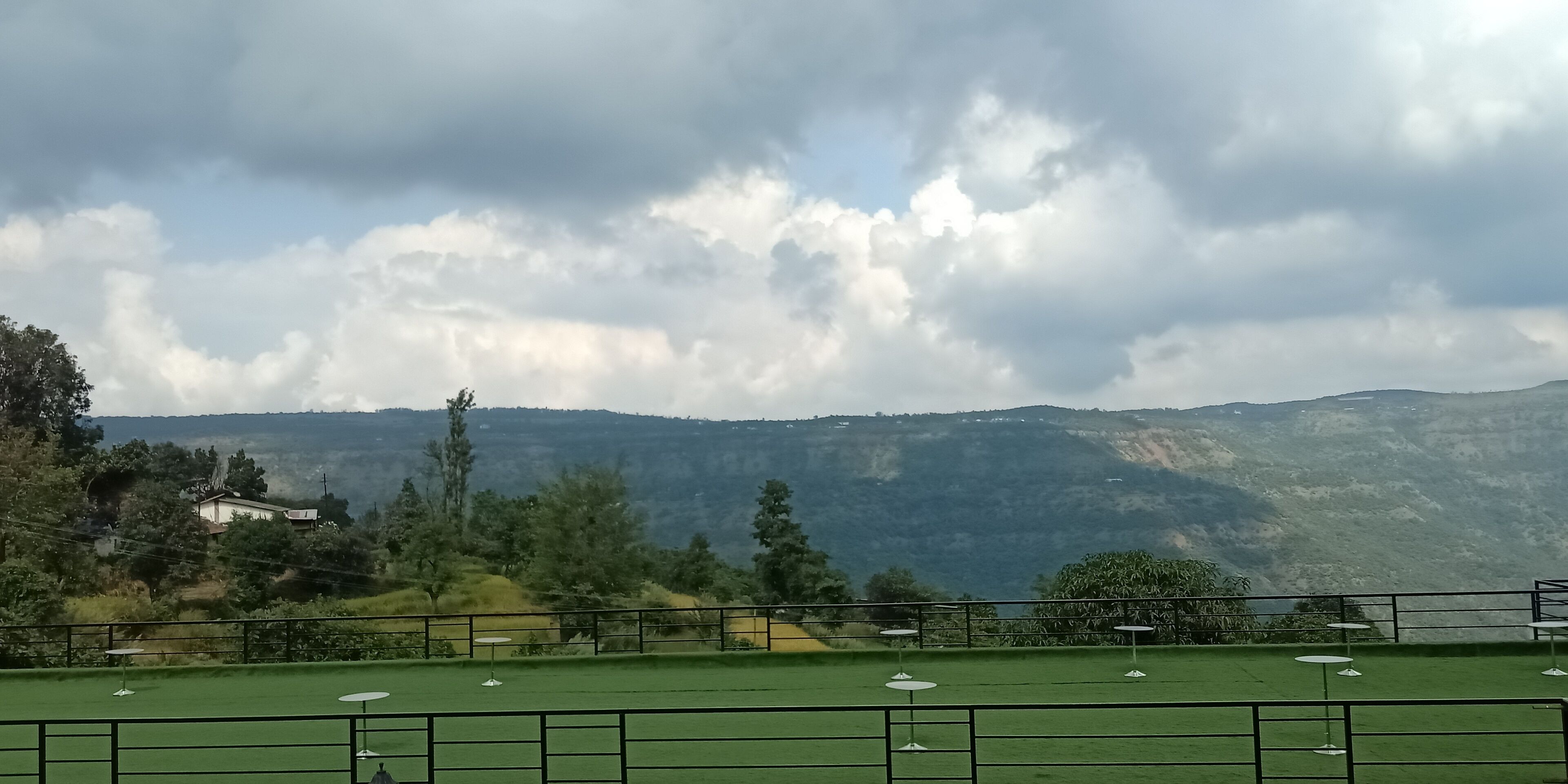 Vista de un césped verde con mesas y sillas blancas, rodeado por una barandilla metálica.